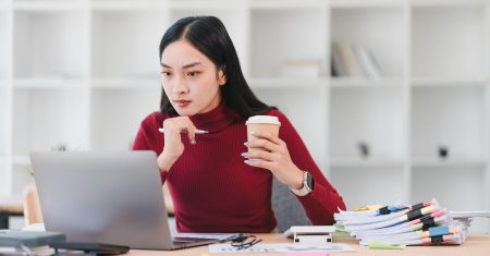 A focused woman in red sweater works on laptop while holding coffee cup in modern office