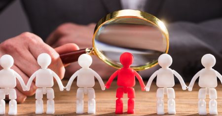Close-up Of A Businessperson's Hand Holding Magnifying Glass Over Red Human Figure On Wooden Desk
